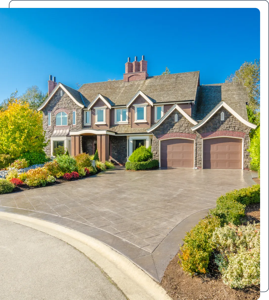 Large stone house with two chimneys, two garages, and a paved driveway, surrounded by trees and landscaped shrubs under a clear blue sky.