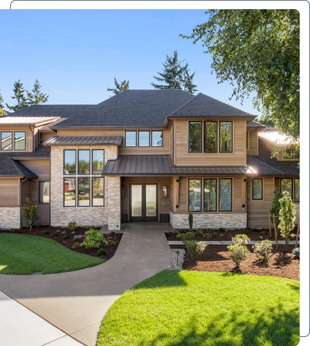 Modern two-story house with a stone and wood facade, large windows, shrubs, and a concrete walkway leading to the entrance, set against a backdrop of trees and a clear blue sky.